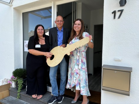 Zwei Frauen und ein Mann stehen mit einem großen, symbolischen Holzschlüssel in der Eingangstür eines Wohngruppenhauses